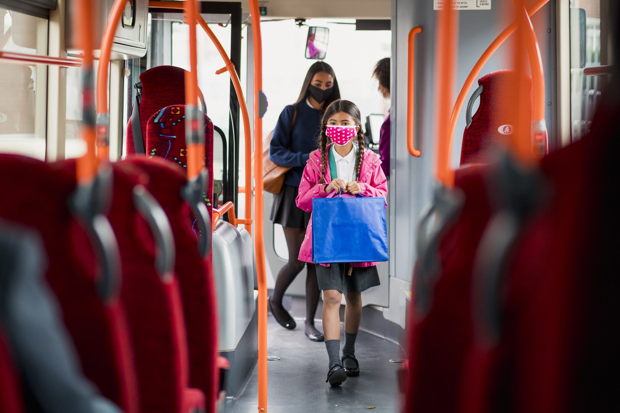 sisters on a public bus wearing masks on their way to school. They are looking for a seat and are wearing their school uniforms.