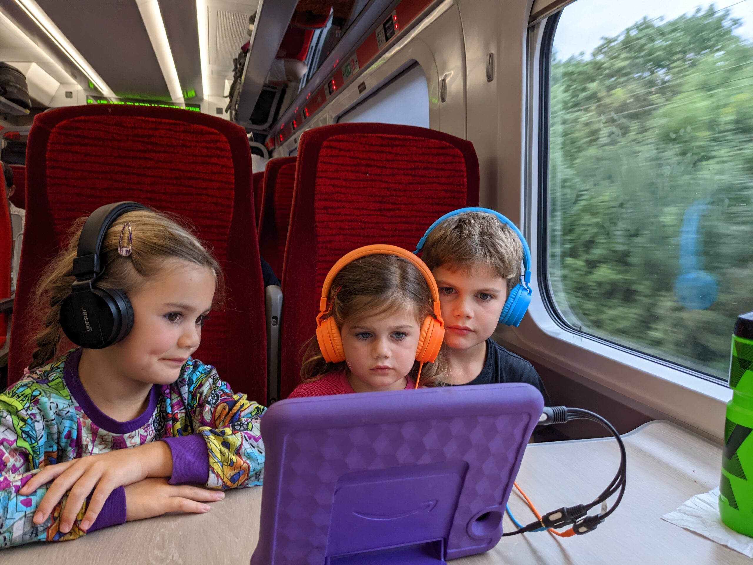 three children on train seat wearing headphones watching a tablet
